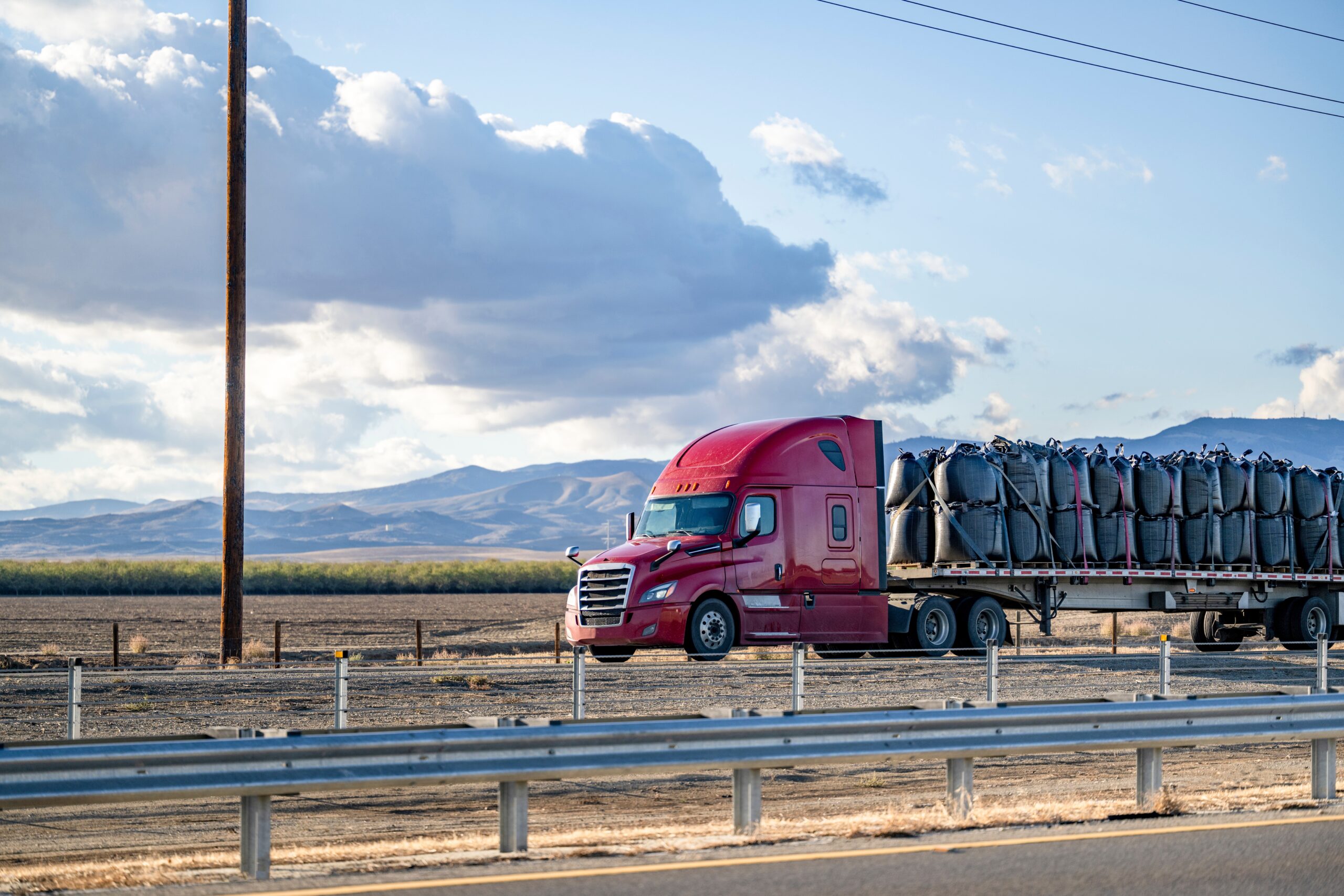 Red semi-truck hauling a flatbed trailer loaded with large bags along a rural highway with mountains in the background.